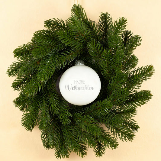Weiß glänzende Weihnachtskugel mit Gravur Frohe Weihnachten im Türkranz auf hellem Hintergrund