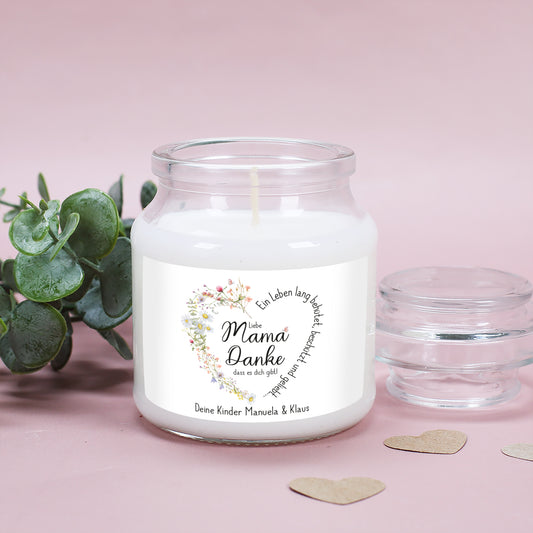 Duftkerze im stabilen, wiederverwendbaren Glas mit Deckel, platziert auf einer sanften rosa Unterlage mit grünen Eukalyptusblättern. Das Etikett zeigt ein florales Herz aus feinen Wiesenblumen mit dem Spruch „Ein Leben lang behütet, beschützt und geliebt…“. In der Mitte steht „Liebe Mama, Danke, dass es dich gibt!“. Die untere Textzeile ist personalisiert mit „Deine Kinder Manuela & Klaus“. Ein persönliches und stilvolles Geschenk für Mütter.
