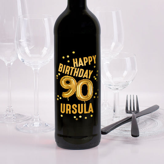 Rotweinflasche mit goldenem „Happy Birthday 90 Ursula“-Aufdruck und Ballon-Design, im Hintergrund Gläser und Besteck