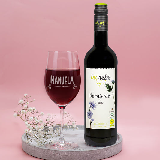 Nahaufnahme von Biorebe Dornfelder Rotwein 2023 mit graviertem Weinglas, Name Manuela, dekoriert mit weißen Blüten auf grauem Tablett.