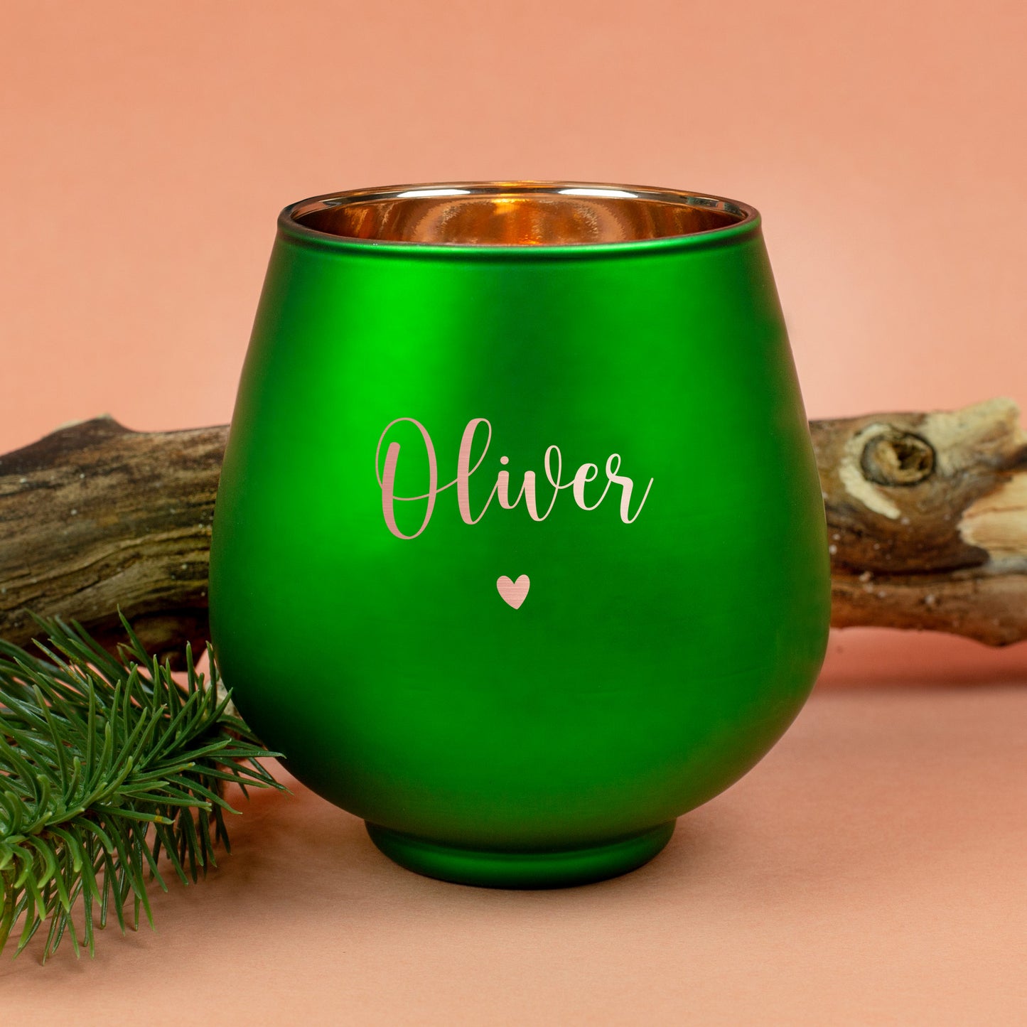 Grünes Windlicht mit Gravur „Oliver“ und Herzmotiv, dekorativ vor Holzstück und Tannenzweig auf orangefarbenem Untergrund.