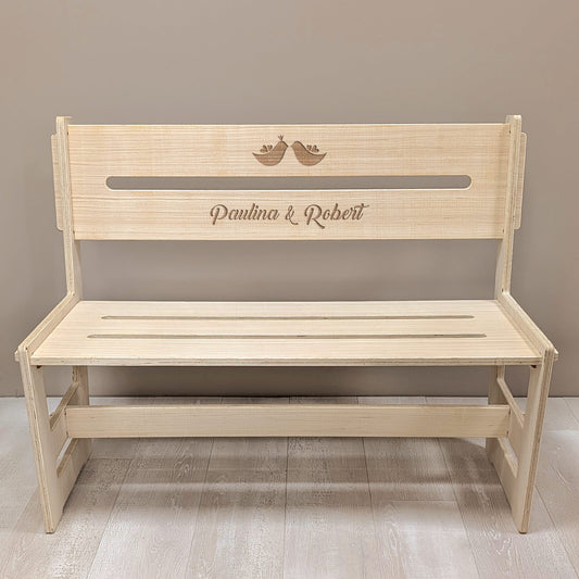 Frontalansicht der gravierten Holzbank mit Vogelmotiv und den Namen "Paulina & Robert" vor heller Wand