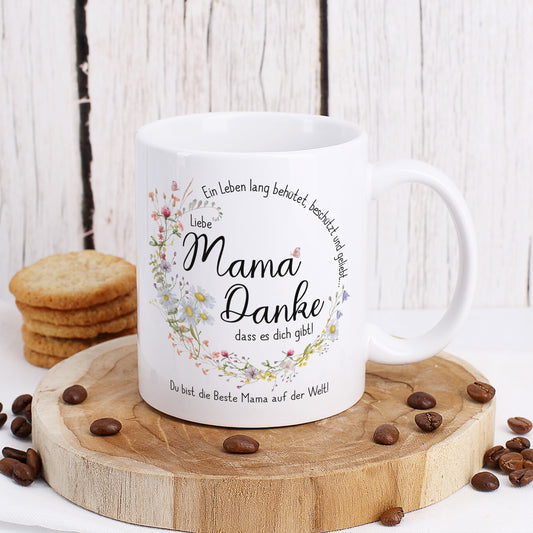Weiße Keramiktasse mit seidenglänzendem Finish, bedruckt mit einem kreisförmigen Wiesenblumen-Kranz und dem Spruch „Ein Leben lang behütet, beschützt und geliebt…“. In der Mitte steht „Liebe Mama, danke, dass es dich gibt!“, ergänzt durch einen personalisierten Wunschtext. Die Tasse steht auf einem Holzbrett, umgeben von Kaffeebohnen und Keksen. Perfektes Geschenk für Mütter zum Muttertag oder Geburtstag.
