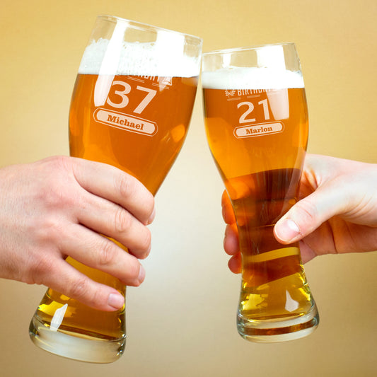 Zwei Hände stoßen mit gefüllten gravierten Weizengläsern an. Links ist das Glas mit der Zahl 37 und dem Namen „Michael“ zu sehen, rechts das Glas mit der Zahl 21 und dem Namen „Marlon“.