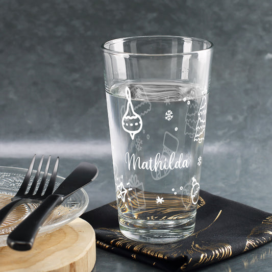 Personalisiertes Glas mit weißen Weihnachtsornamenten, Name Mathilda, gefüllt mit Wasser, auf rustikalem Tisch mit Besteck.