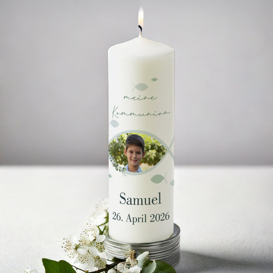 Weiße Kerze zur Kommunion mit Foto. Oben steht „meine Kommunion“, darunter das Foto eines Jungen, eingerahmt von einem stilisierten Fischsymbol. Darunter Name „Samuel“ und Datum „26. April 2026“. Die Kerze ist angezündet und von weißen Blüten umgeben.