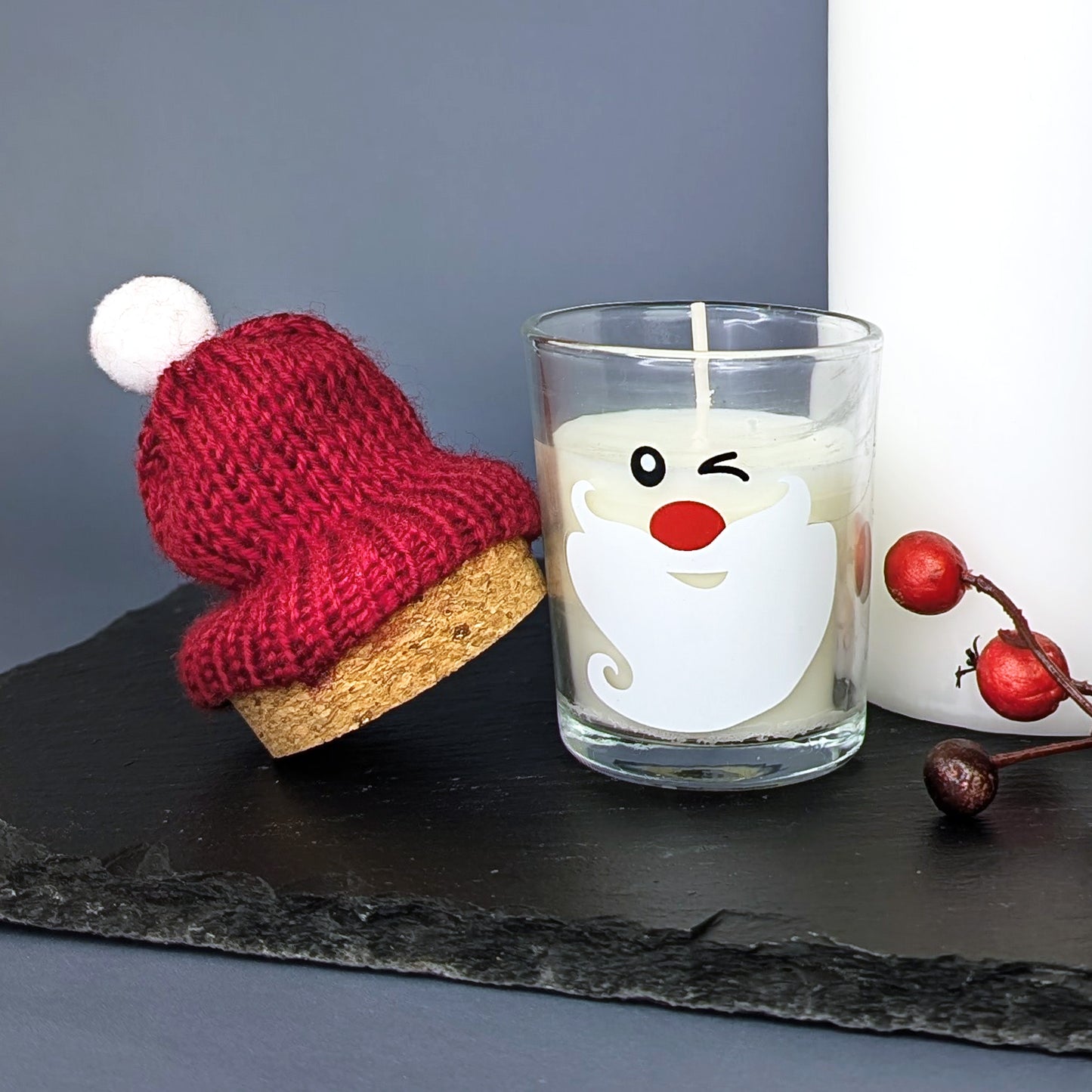Kerzenglas mit abgenommener Mütze – weiße Kerze mit Weihnachtsgesicht und rote Pudelmütze daneben, stimmungsvolle Deko mit Beerenzweig.