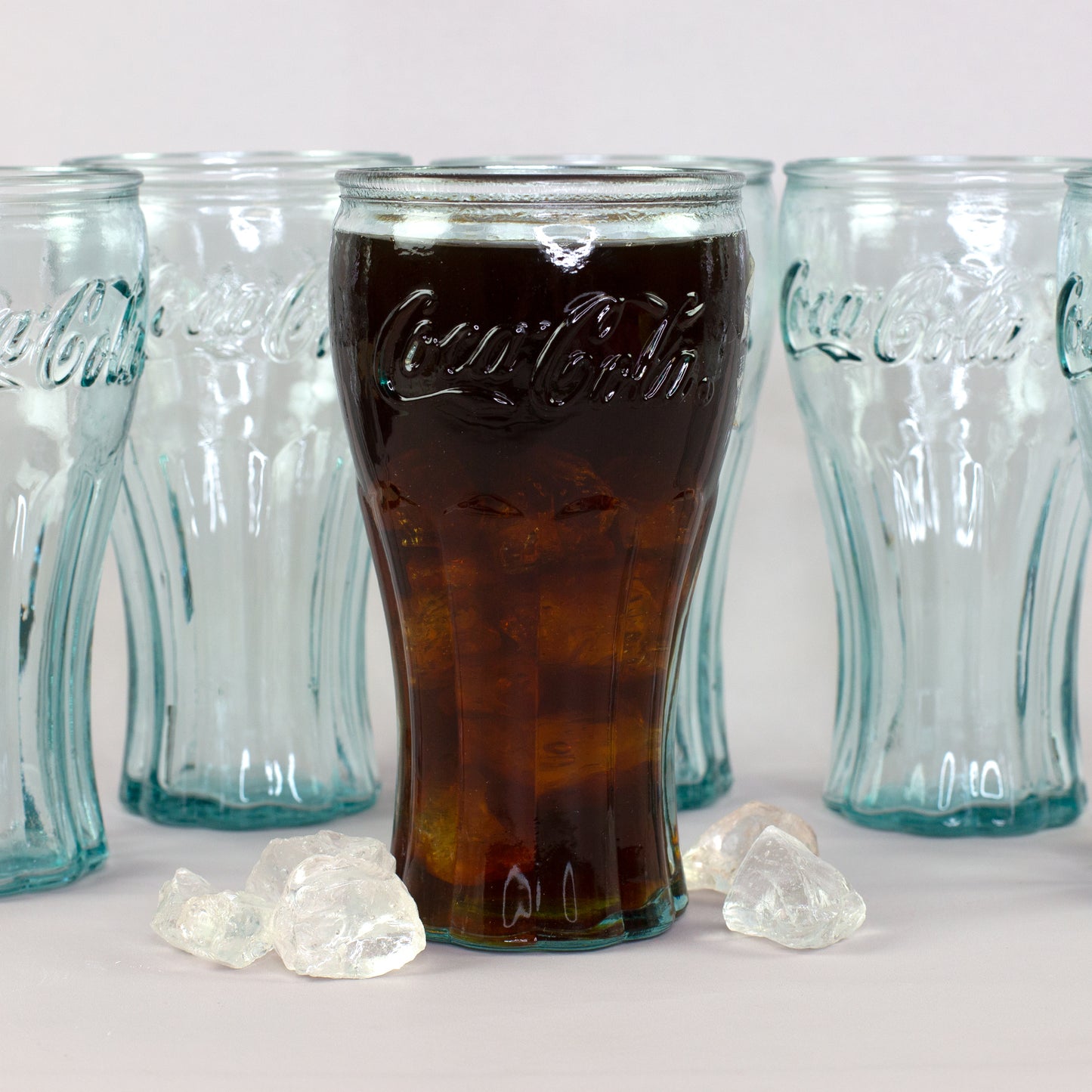 Retro Coca-Cola Trinkglas mit Cola und Eiswürfeln vor mehreren Gläsern aus recyceltem Glas