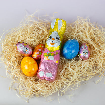 Riegelein Oster-Mischbeutel 100g ausgepackt liegend auf ausgebreiteter Holzwolle zusammen mit zwei gefärbten Eiern