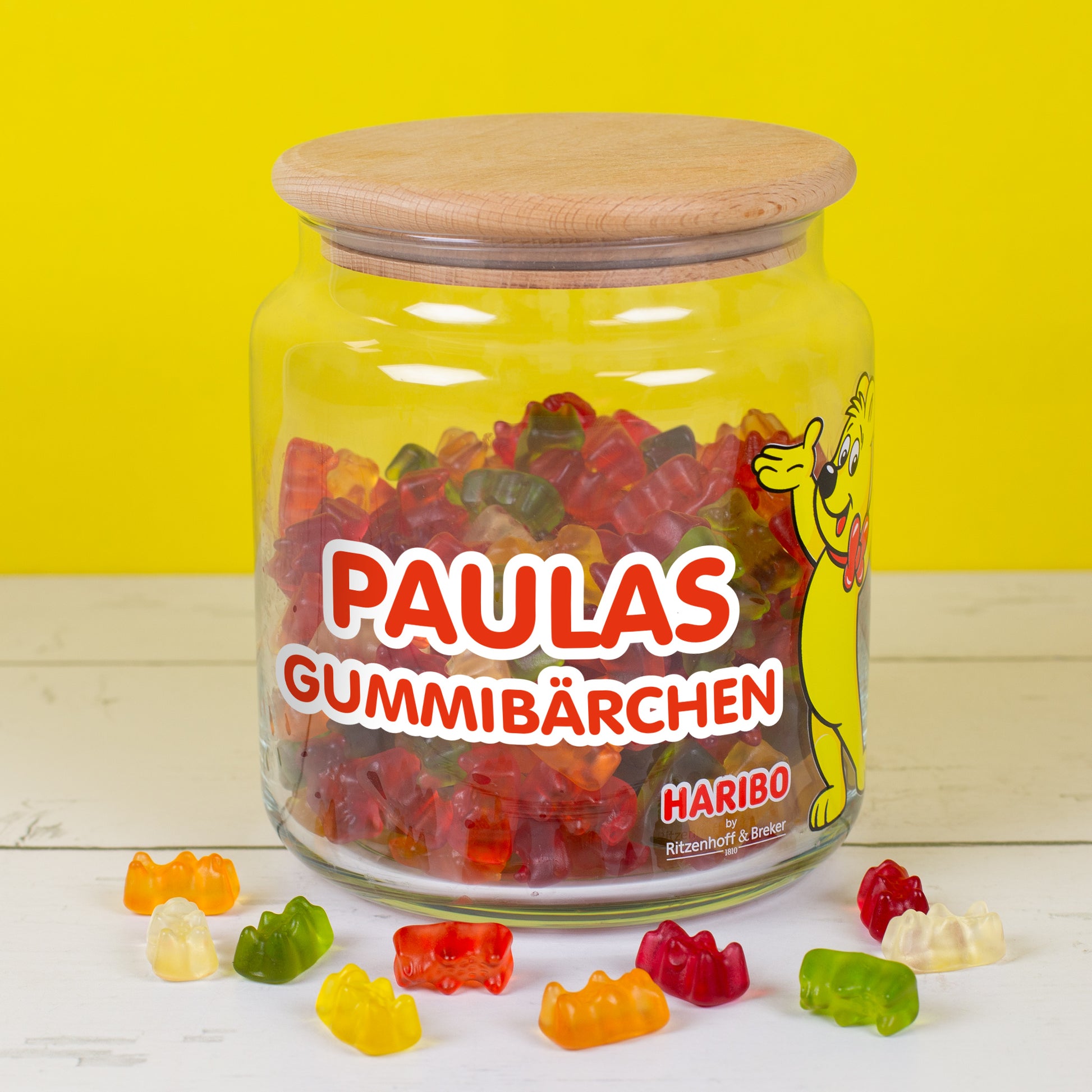 HARIBO Glasdose 1,1 l mit personalisiertem rotem Aufdruck ‚Paulas Gummibärchen‘, Goldbär-Motiv, bunt gefüllt, auf gelbem Hintergrund.