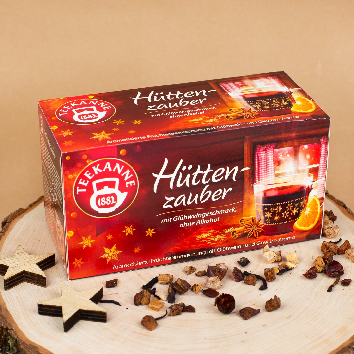Packung „Teekanne Hüttenzauber“ auf Holzscheibe platziert, dekoriert mit Sternen, Gewürzen und losen Teestückchen.