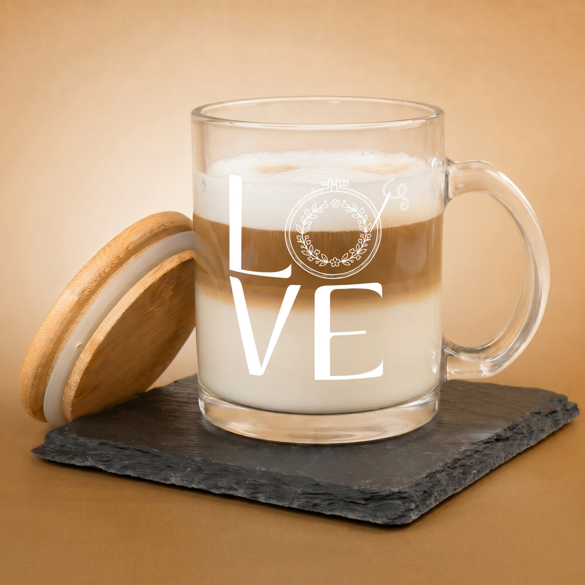 Glastasse mit weißem LOVE Stickmotiv, Kaffee mit Milch und danebenstehendem Bambusdeckel auf Schieferplatte