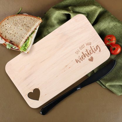 Holzbrettchen mit Gravur „Du bist mir wichtelig“ und Herzausschnitt, dekoriert mit Sandwich und Tomaten.
