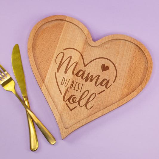 Herzförmiges Holzbrettchen mit Gravur Mama du bist toll neben goldenem Besteck auf lila Hintergrund
