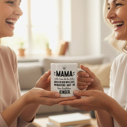 Zwei Frauen überreichen sich lachend eine weiße Keramiktasse mit Mama-Spruch als Geschenk in wohnlicher Umgebung