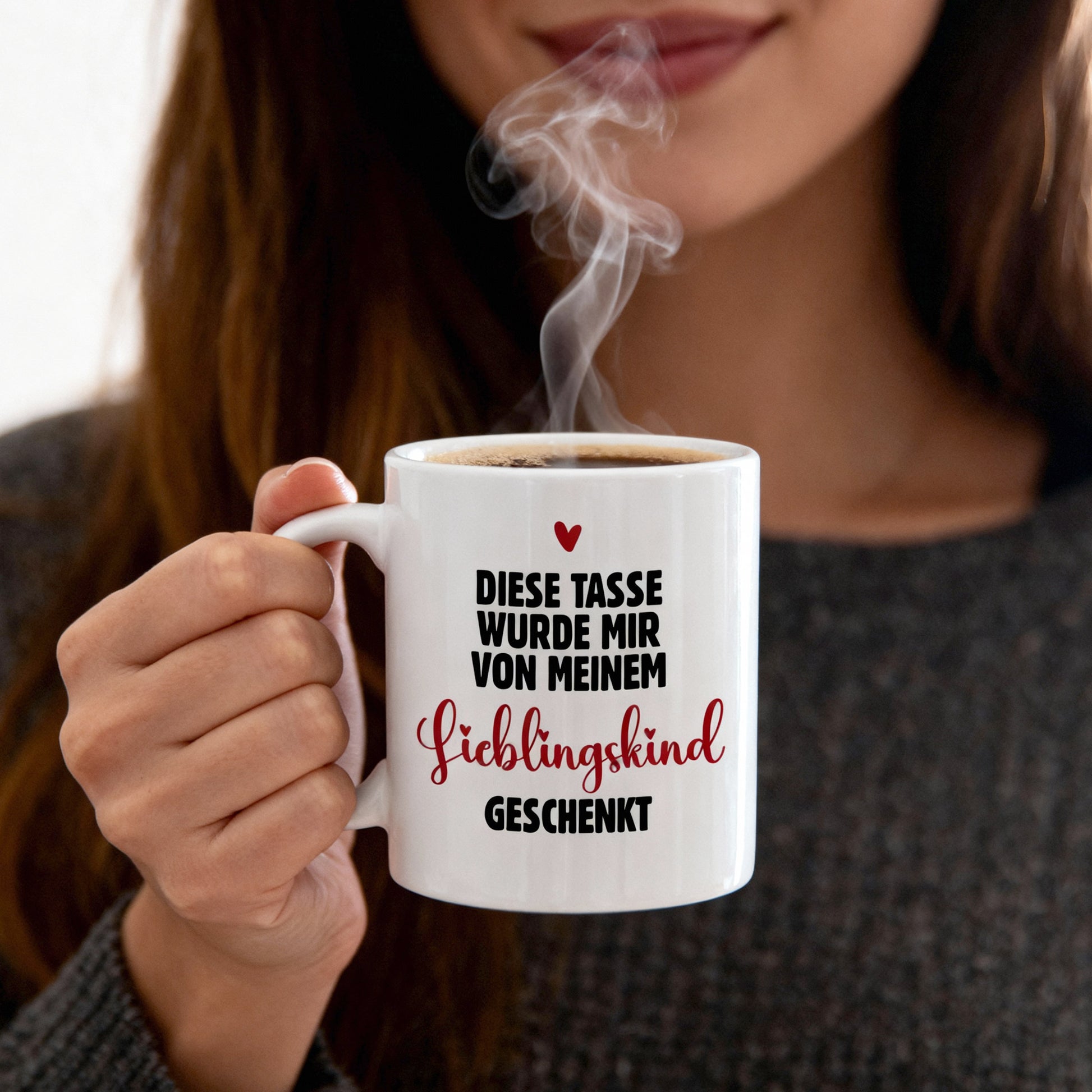 Weiße Keramiktasse mit dem Spruch „Diese Tasse wurde mir von meinem Lieblingskind geschenkt“, gehalten in der Hand, mit aufsteigendem Dampf