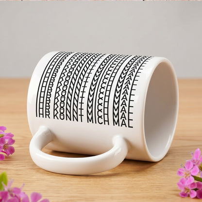 Seitlich liegende Tasse mit verstecktem Schriftzug „IHR KÖNNT MICH MAL“ neben kleinen Blumen