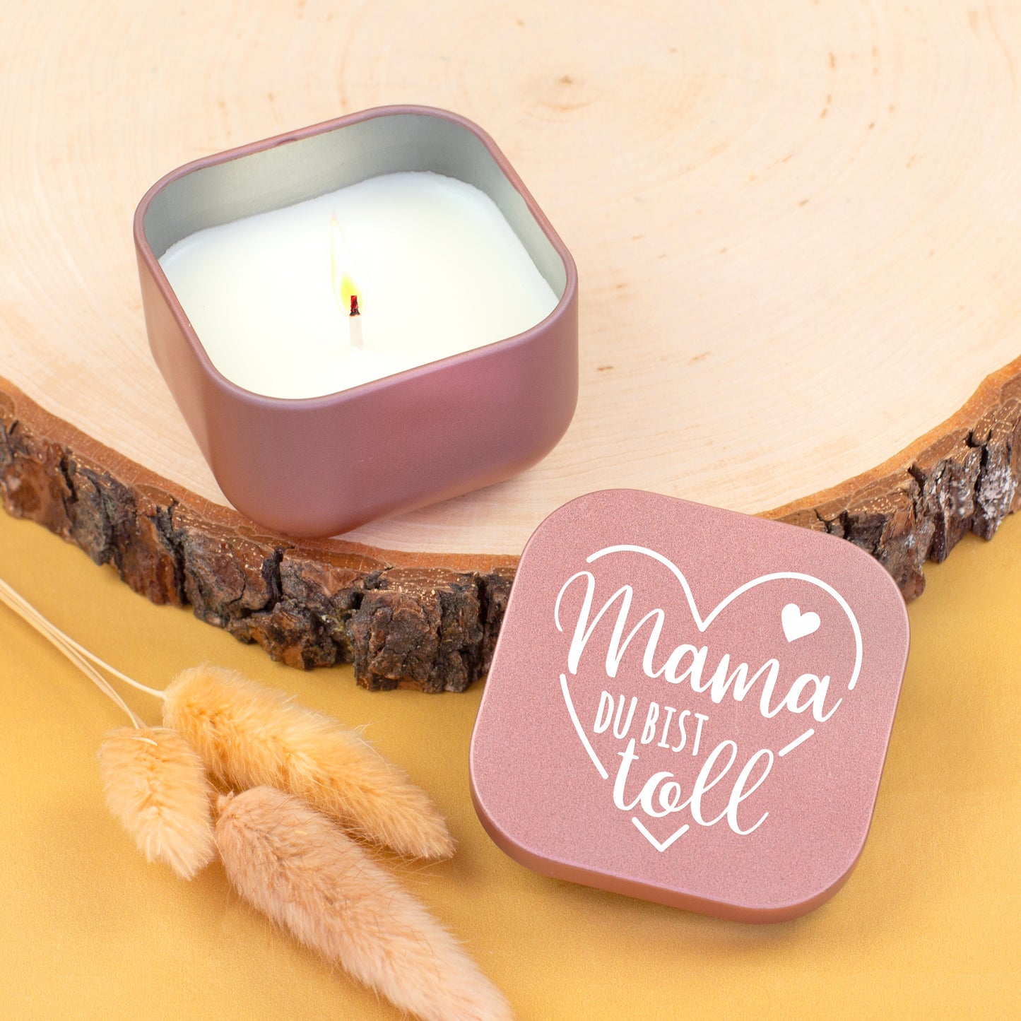 Roséfarbene Duftkerze „Mama du bist toll“ geöffnet mit brennender Flamme auf Holzscheibe