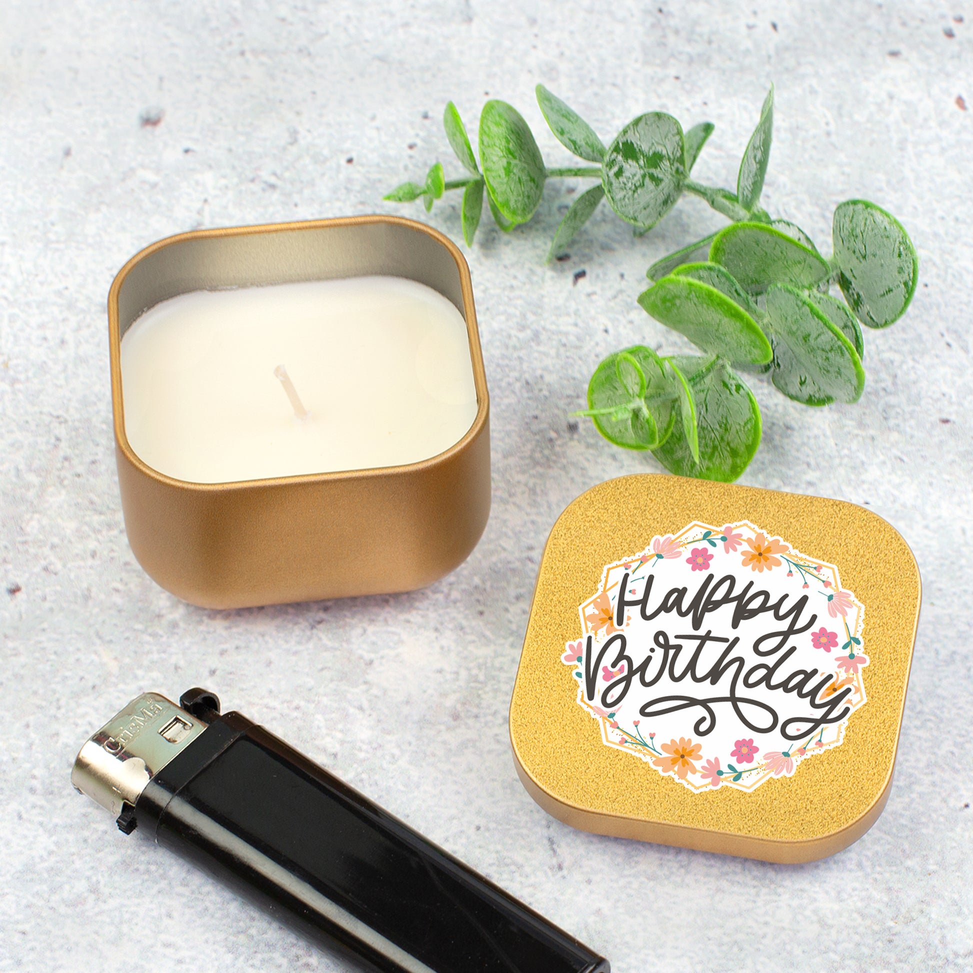 Goldene Duftkerze mit Happy Birthday Motiv neben Feuerzeug und Eukalyptuszweig