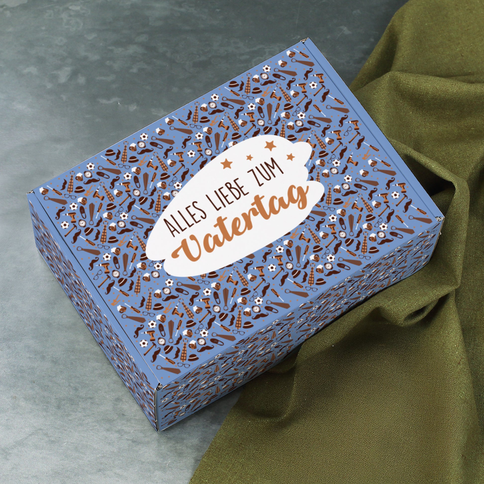 Geschenkverpackung zum Vatertag auf grauem Untergrund, seitlich geöffnet mit Olivstoff im Hintergrund.