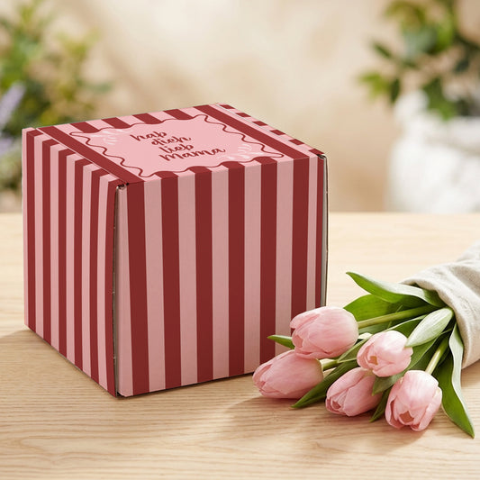 Geschenkbox zum Muttertag mit rosa-rotem Streifenmuster und Spruch „hab dich lieb Mama“ auf hellem Holztisch neben rosa Tulpen