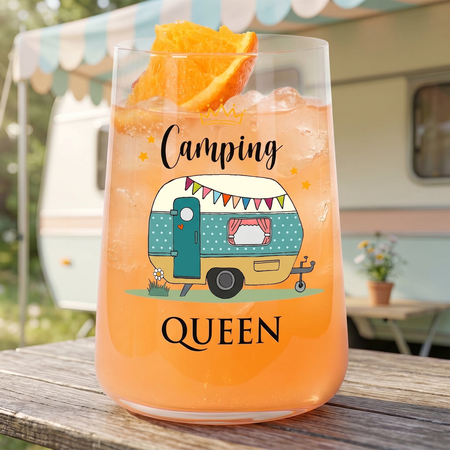 Trinkglas mit Camping Queen Wohnwagen-Motiv vor Wohnwagen-Hintergrund und orangefarbenem Getränk