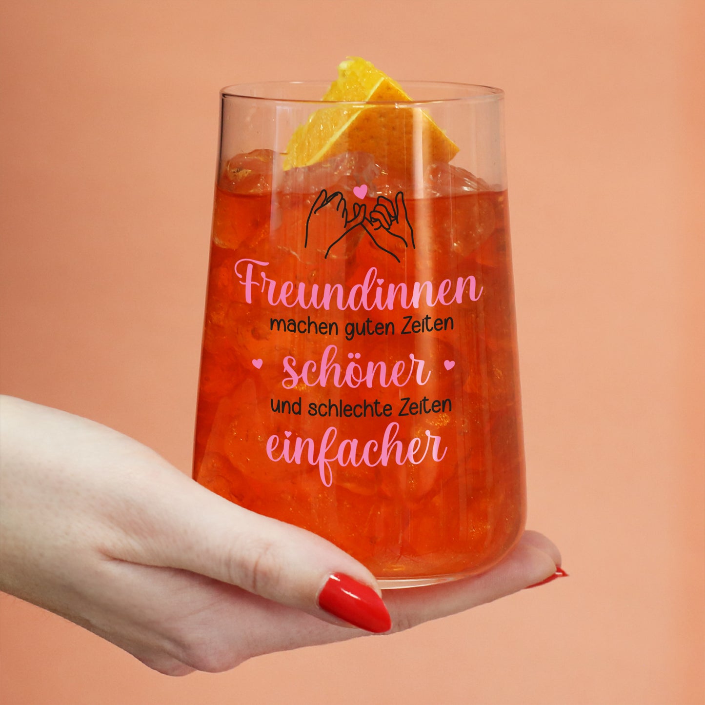 Trinkglas mit rotem Getränk, Eiswürfeln, Orangenscheibe und Freundinnen-Motiv in der Hand gehalten