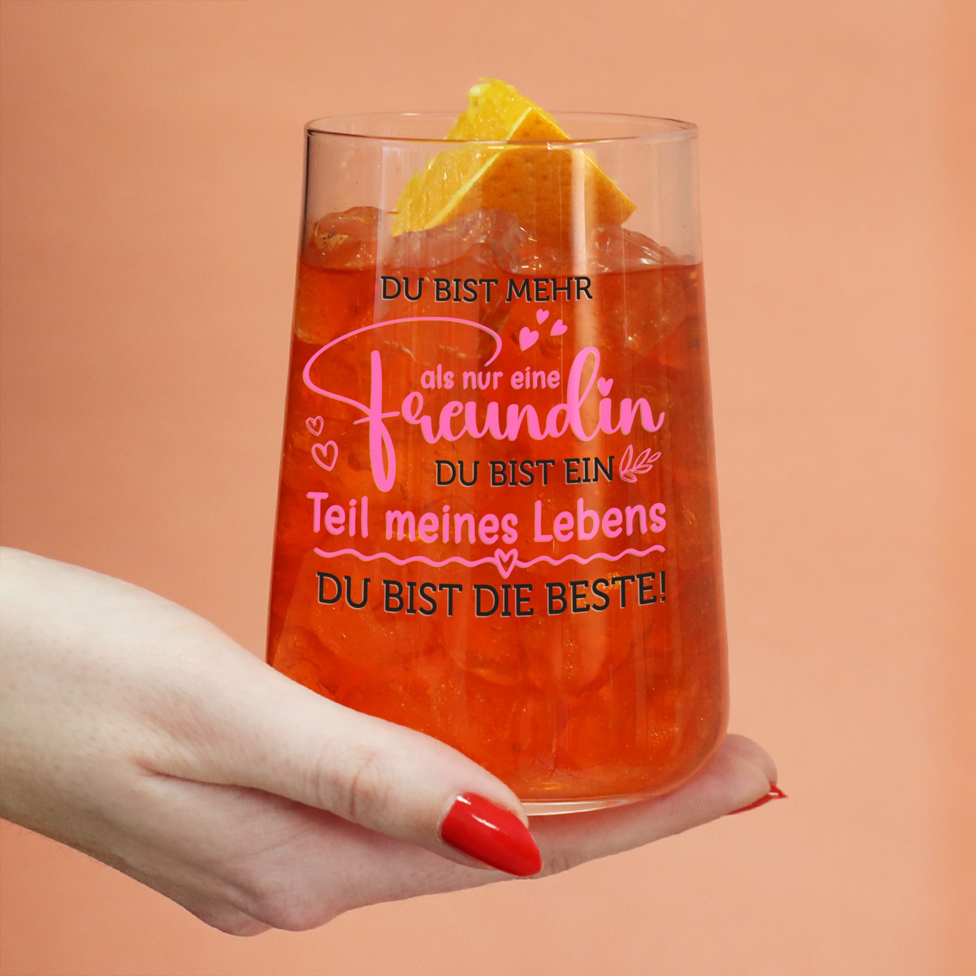 Spritzglas mit Freundinnen-Spruch und orangefarbenem Getränk, gehalten in einer Hand vor farbigem Hintergrund