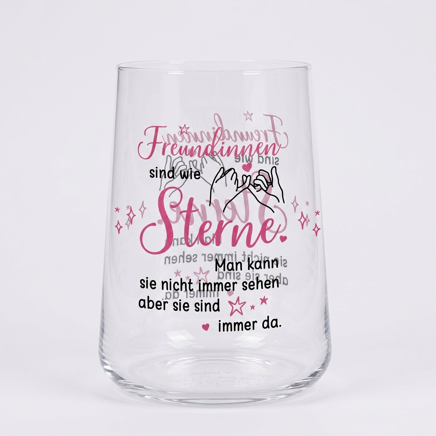Leeres Trinkglas mit rosa-schwarzem Motivdruck „Freundinnen sind wie Sterne“ und Illustration zweier Hände auf weißem Hintergrund