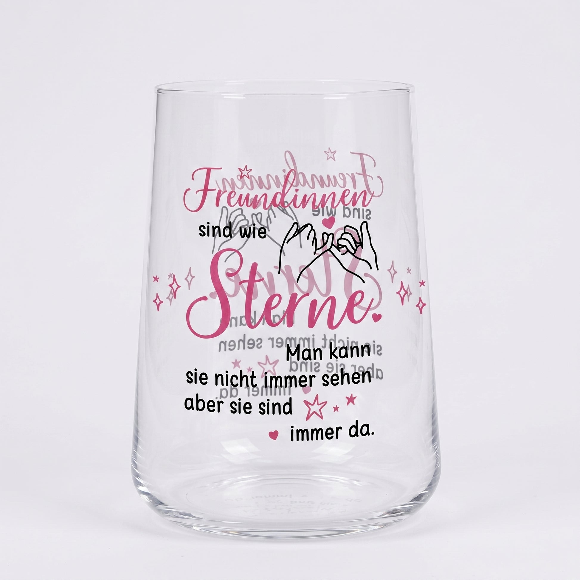 Leeres Trinkglas mit rosa-schwarzem Motivdruck „Freundinnen sind wie Sterne“ und Illustration zweier Hände auf weißem Hintergrund