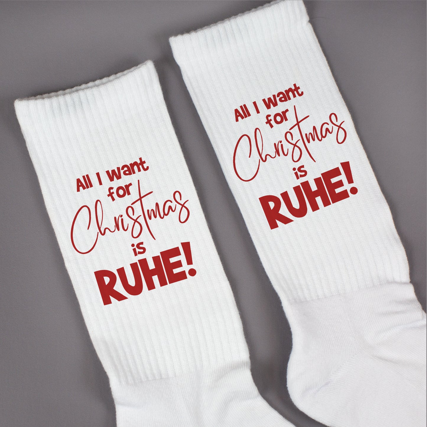 Paar weiße Tennissocken mit rotem Weihnachtsdruck ‚All I want for Christmas is RUHE!‘ flach nebeneinander liegend.