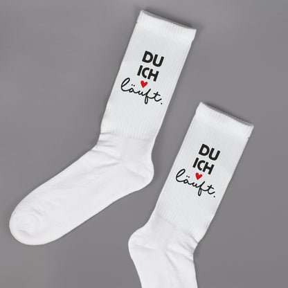 Zwei weiße Socken liegend auf grauem Hintergrund, Aufdruck „DU ICH läuft.“ mit Herz gut sichtbar