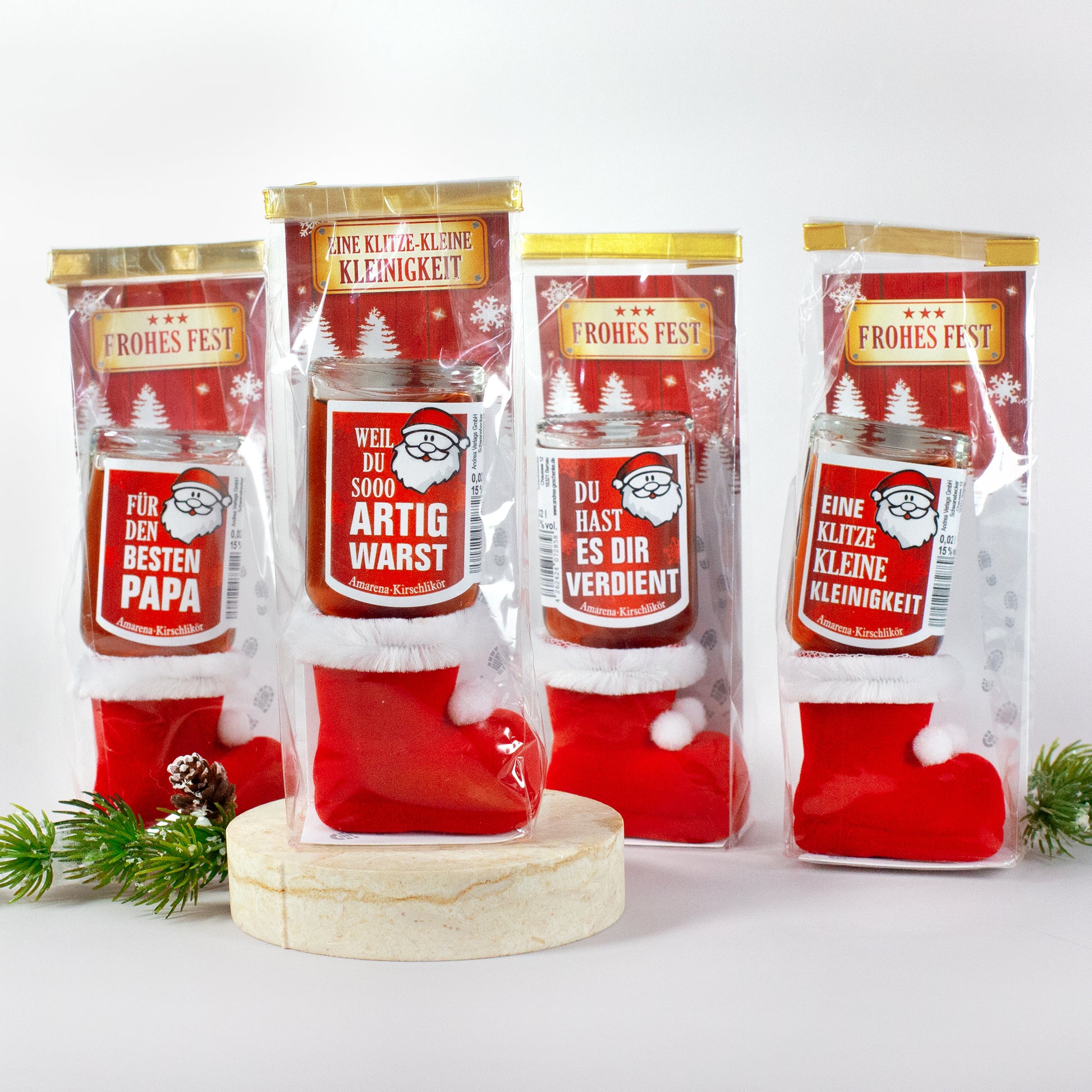 Übersicht aller vier Varianten in der Klarsichtverpackung. Likörflaschen mit unterschiedlichen Sprüchen stehen auf einer hellen Präsentationsfläche, umgeben von Tannenzweigen. Ideal zur Darstellung aller Spruchoptionen.