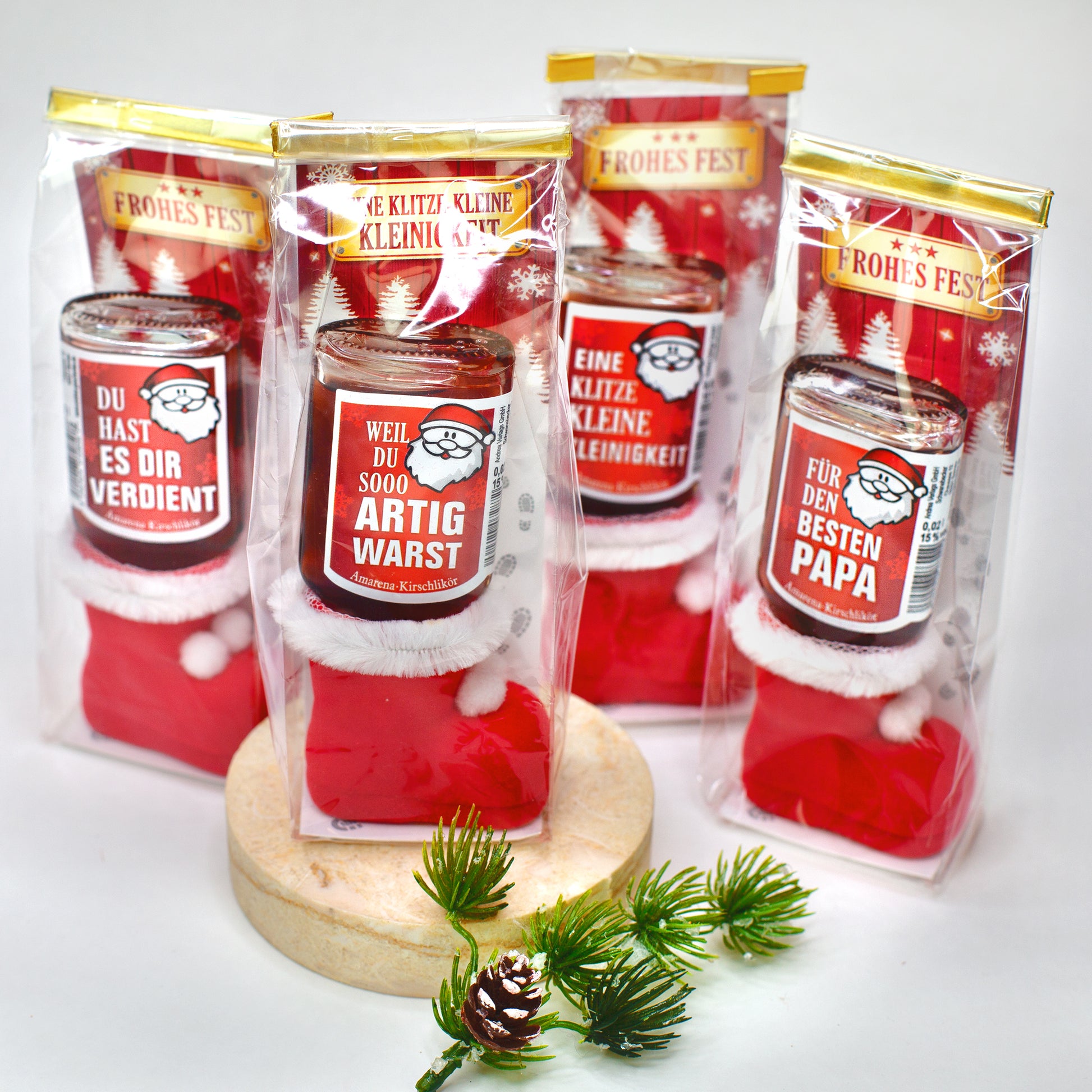 Vier rote Nikolausstiefel mit je einer auf dem Kopf stehenden Likörflasche, verpackt in Klarsichtbeuteln mit Goldverschluss. Jeder Likör trägt ein anderes Etikett mit Sprüchen wie „Artig warst“ oder „Bester Papa“. Im Hintergrund Weihnachtsmotive und Dekozweig.