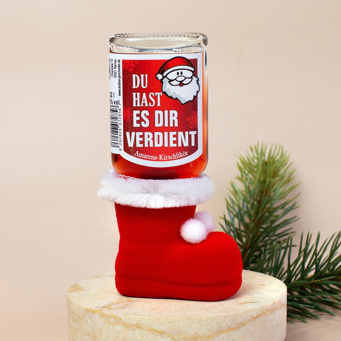 Flasche „Du hast es dir verdient“ aufrecht im Stiefel platziert. Der Stiefel steht auf einem hellen Podest, im Hintergrund ein kleiner Zweig. Fokus liegt auf der Etikettenansicht und Stiefel.