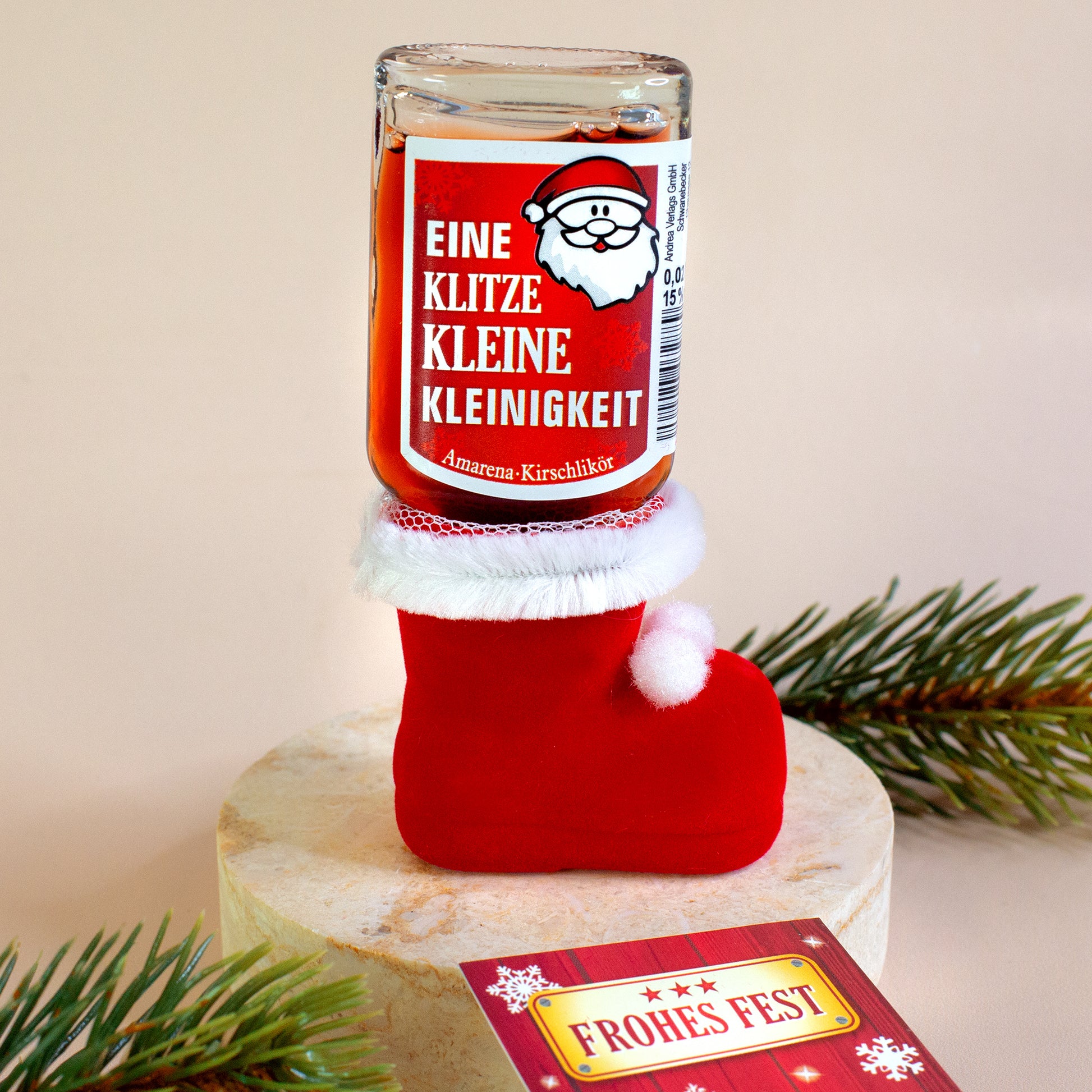 Flasche mit „Eine klitzekleine Kleinigkeit“ steht im roten Nikolausstiefel auf hellem Podest. Rechts im Bild eine Weihnachtskarte „Frohes Fest“, rundherum grüne Zweige.