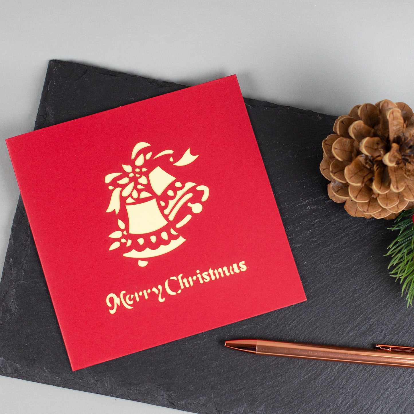 Rote Weihnachtskarte mit goldenem Glockenmotiv und „Merry Christmas“-Schriftzug, dekorativ auf Schieferplatte arrangiert.