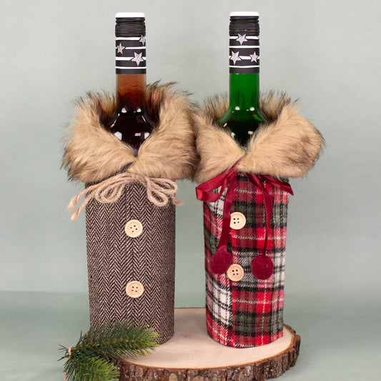 Zwei Flaschen Geschenkverpackungen in Mantel-Optik mit Pelzkragen, links in Braun, rechts kariert in Rot-Grün, auf Holzplatte dekoriert.
