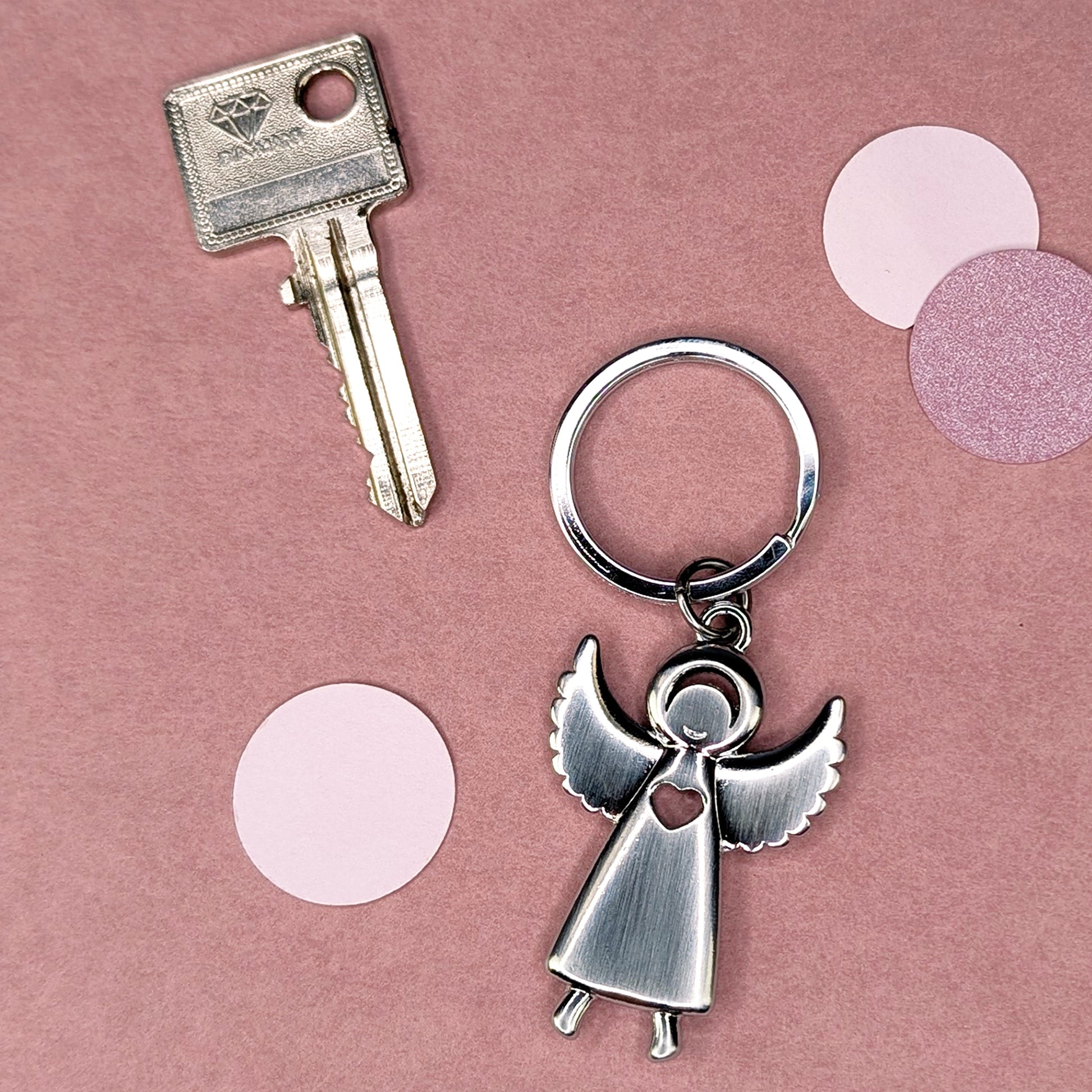 Schlüsselanhänger in Engel-Form neben einem Schlüssel, von oben fotografiert auf rosafarbenem Hintergrund