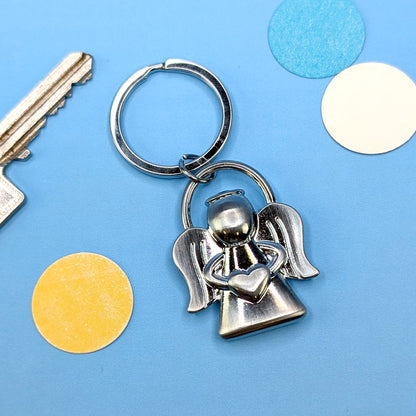 Metallischer Schutzengel-Schlüsselanhänger mit herzförmigem Detail, liegend auf blauem Untergrund mit bunten Kreisen