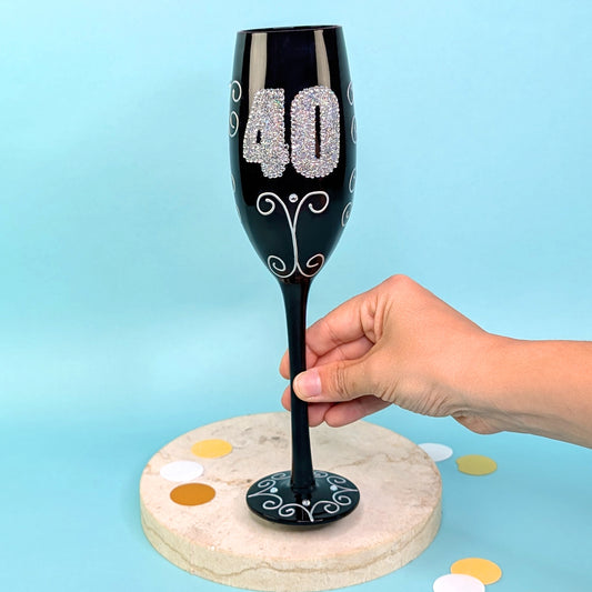 Schwarzes Sektglas mit glitzernder 40 und Ornamenten, frontal fotografiert, gehalten von einer Hand