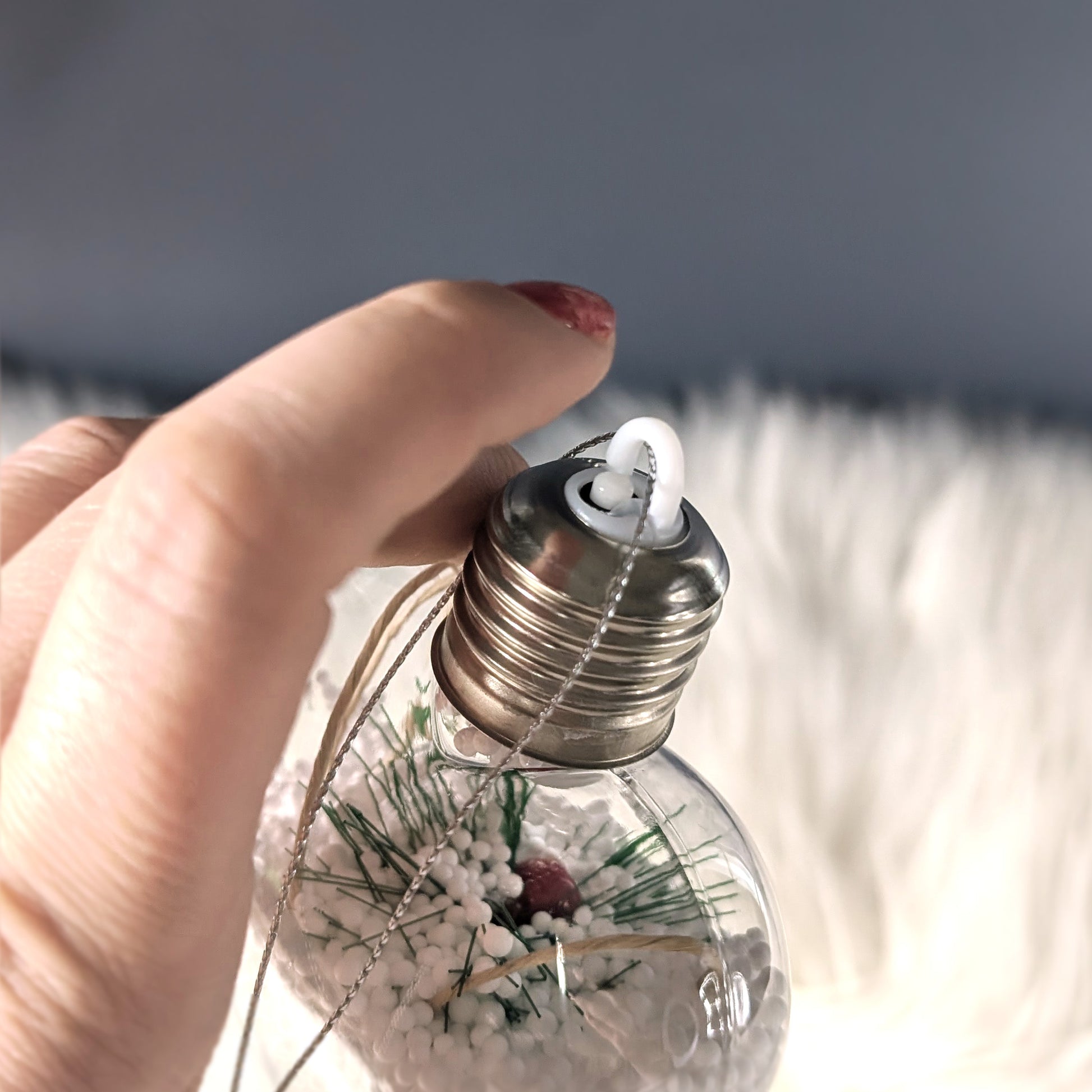 Hand schaltet LED-Weihnachtskugel mit Metallverschluss ein – im Inneren Schneeoptik mit Tannenzweig und roten Beeren.