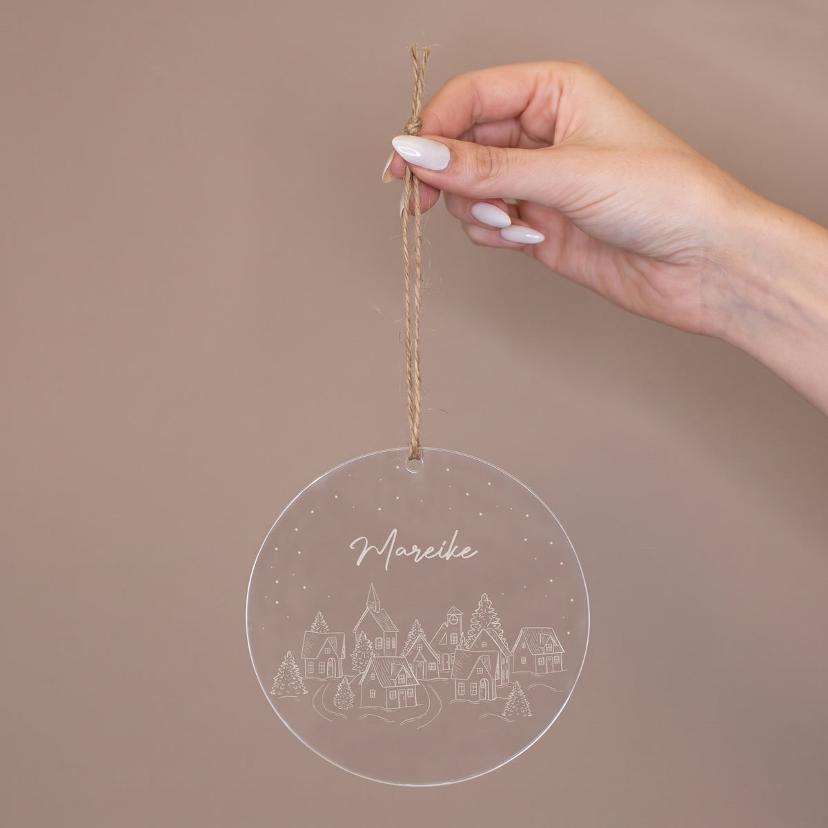 Personalisierte Acryl Weihnachtskugel wird an einem Juteband gehalten und zeigt Wunschtext und Winterdorf