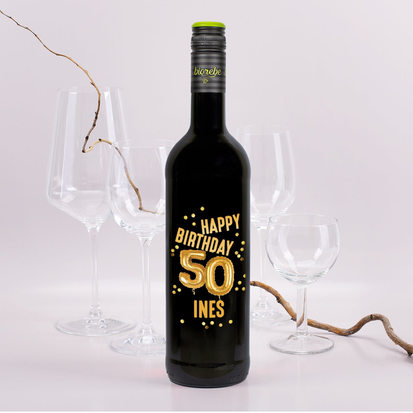 Schwarze Weinflasche mit goldener Ballonzahl „50“ und dem Text „Happy Birthday Ines“ auf dem Etikett, im Hintergrund Weingläser und Rebenzweig