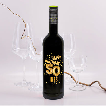 Schwarze Weinflasche mit goldener Ballonzahl „50“ und dem Text „Happy Birthday Ines“ auf dem Etikett, im Hintergrund Weingläser und Rebenzweig