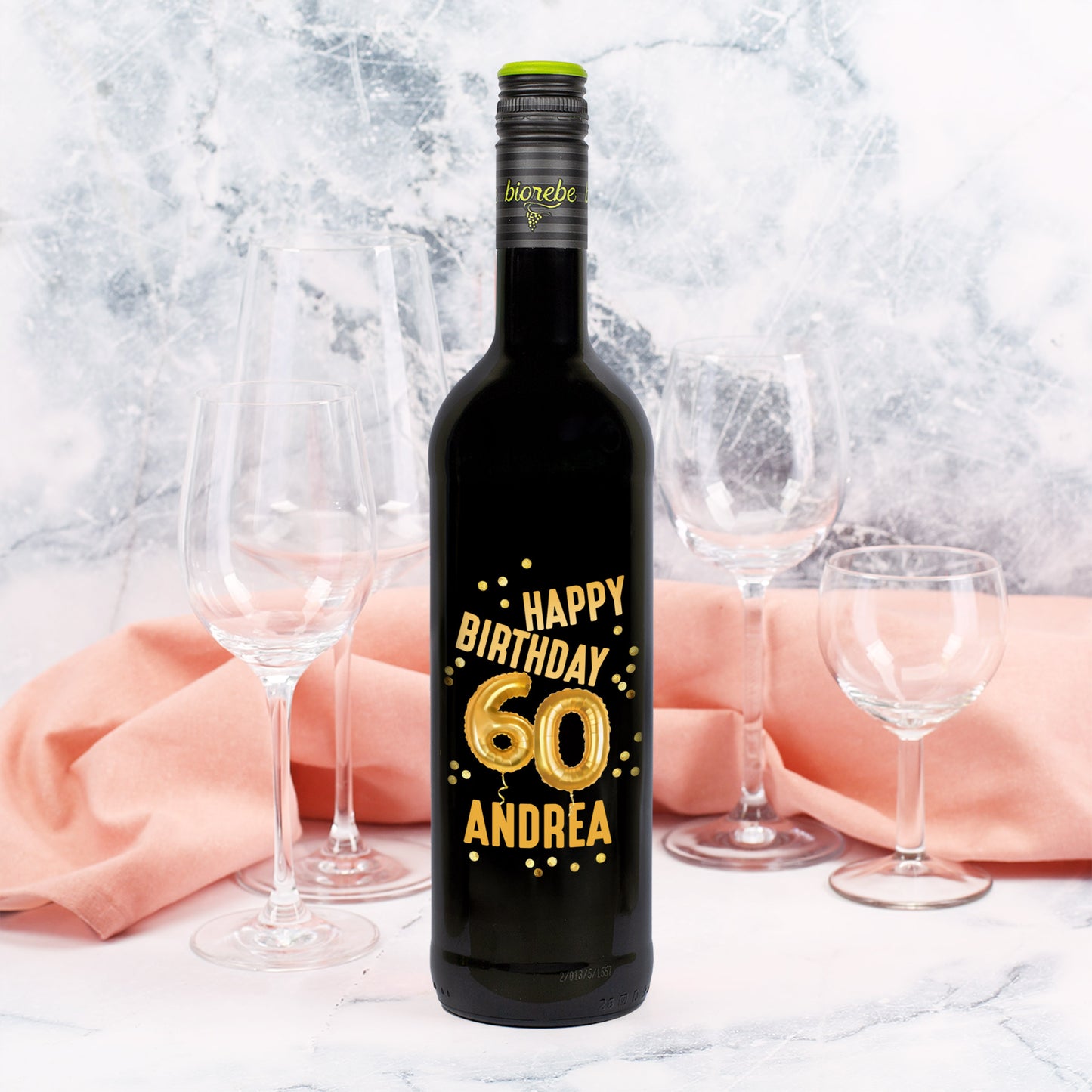 Weinflasche mit schwarzem Etikett zum 60. Geburtstag, goldene Ballons „60“, Name Andrea, Hintergrund mit rosa Stoff und Weingläsern