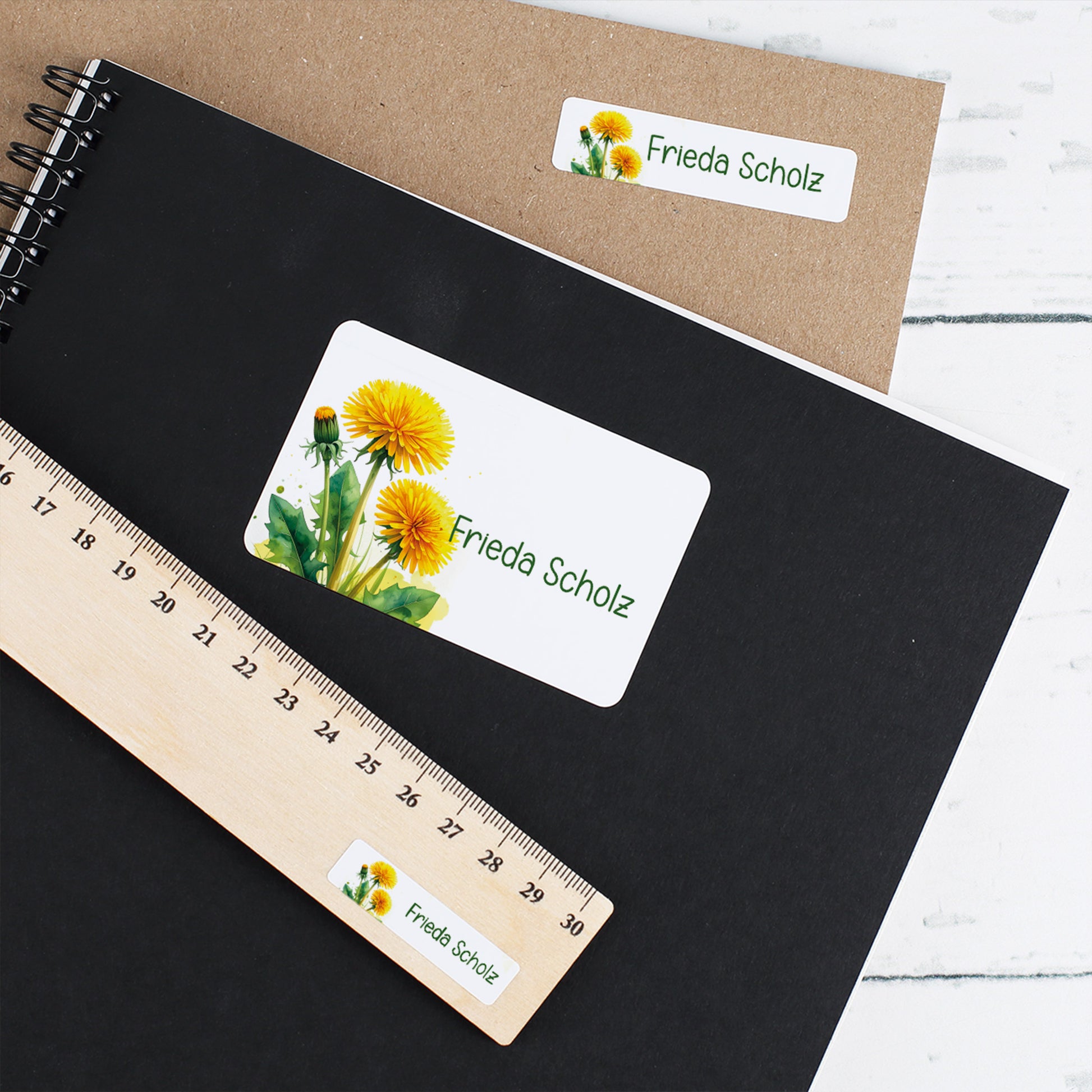 Zwei Schulhefte mit personalisierten Löwenzahn-Aufklebern, Name Frieda Scholz, auf schwarzem und braunem Umschlag