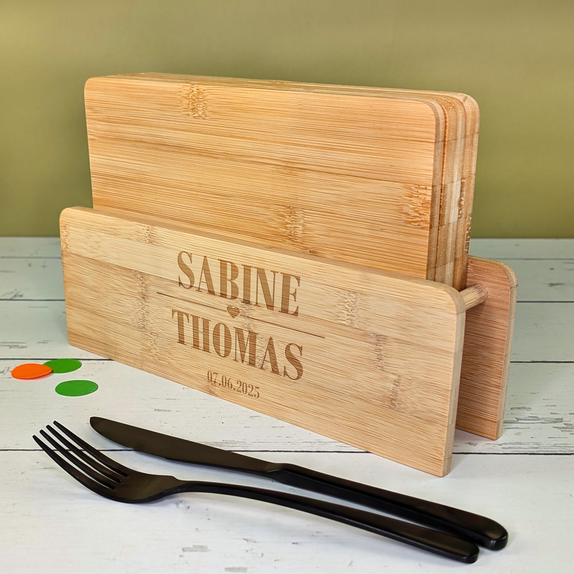 Bambusbrettchen-Set mit Gravur „Sabine & Thomas“ und Datum, daneben schwarzes Besteck auf hellem Tisch
