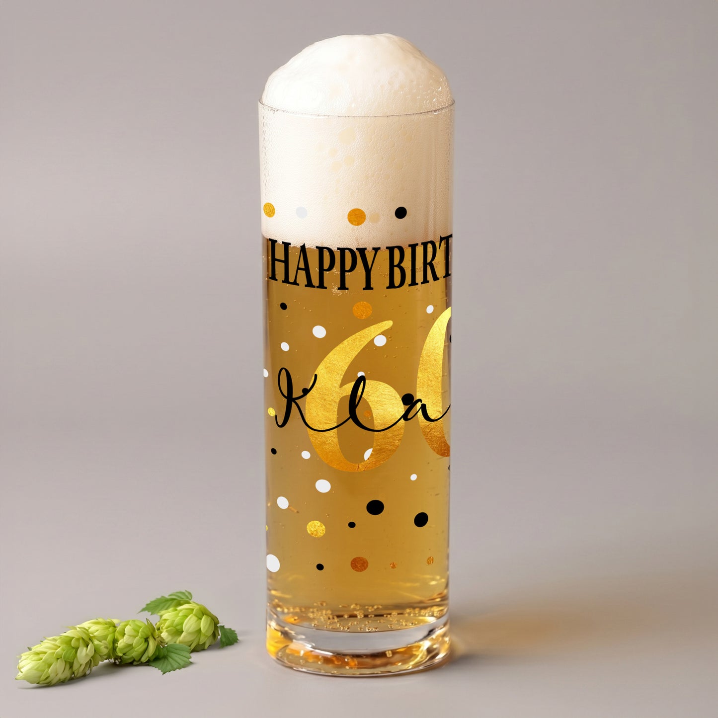 Kölschglas mit Geburtstagsmotiv, goldener Zahl 60 und personalisiertem Namen auf grauem Hintergrund, daneben grüne Hopfendolden.