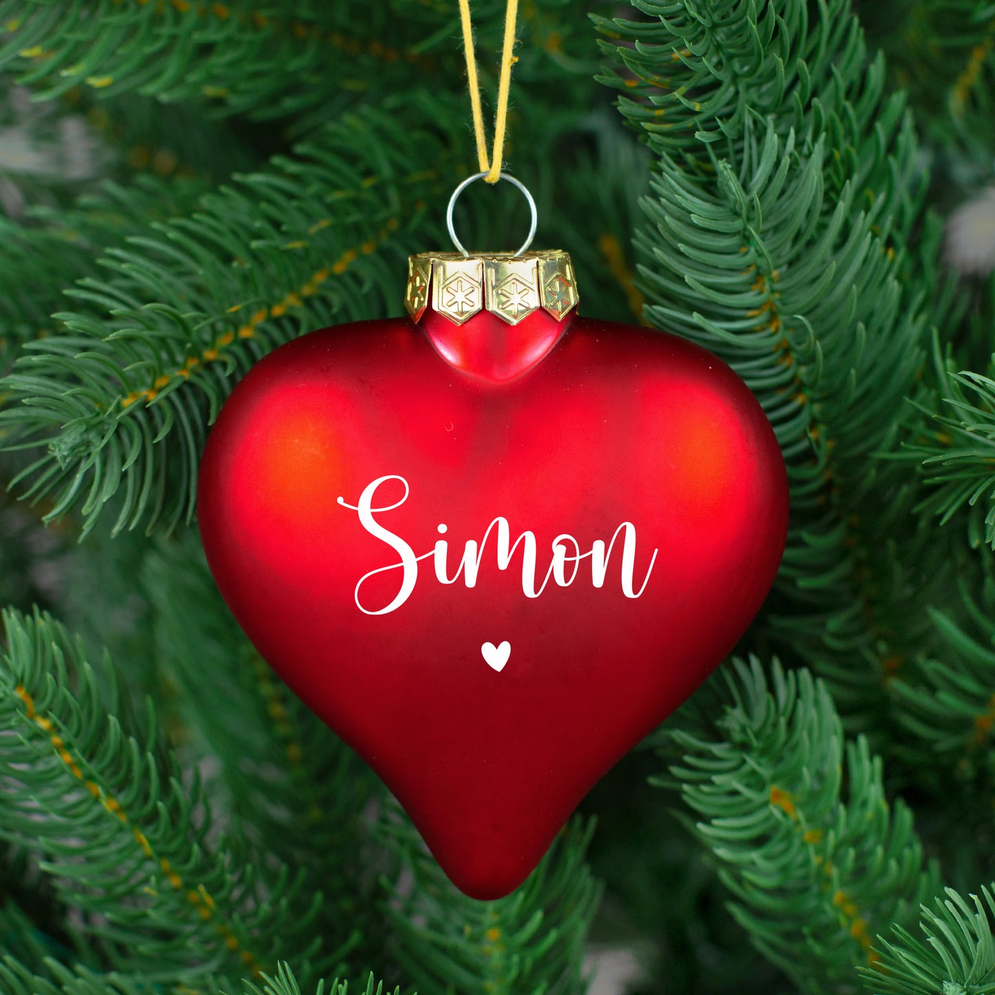 Rote, matte Herz-Christbaumkugel mit weißem Namen „Simon“ und kleinem Herz, an grünem Weihnachtsbaum.